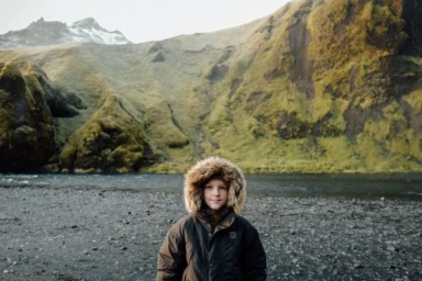 Child wearing a dark parka with a furry hood standing in front of a dark rocky riverbank and grassy green mountains.