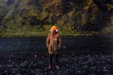 Man wearing a brown winter coat and orange beanie standing on a dark sandy shore near a river, photographed by Christa Renee.
