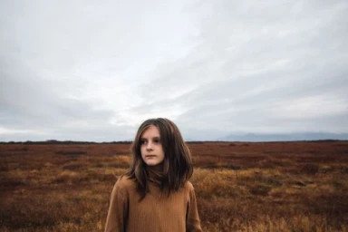 Young girl wearing a brown ribbed turtleneck standing outdoors against a vast barren landscape under a cloudy sky.