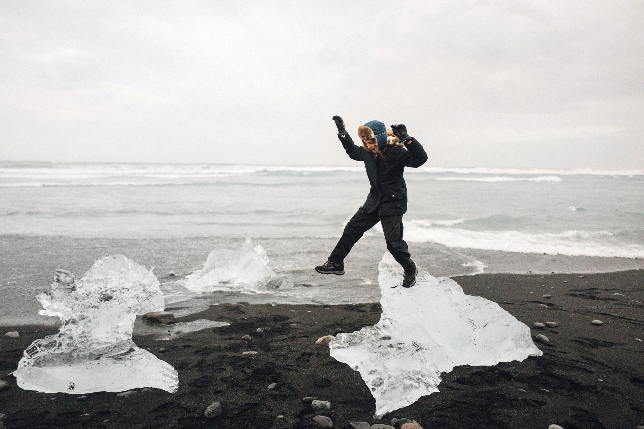 Person jumping between large chunks of glacial ice on a dark volcanic black sand beach with ocean waves behind.