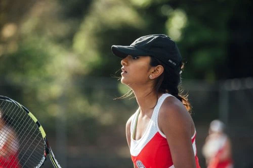 Profile view of a female tennis player wearing a black cap and red and white sports top holding a racket