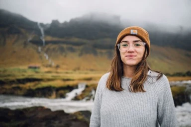 Woman wearing an orange beanie and clear glasses standing in front of a misty mountain landscape and waterfall.