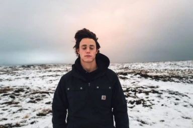 Young man with dark hair in a black winter jacket standing against a snowy landscape under an overcast sky.