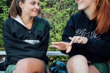 Two young women sitting together outdoors, one wearing a black windbreaker and green skirt and the other a black hoodie