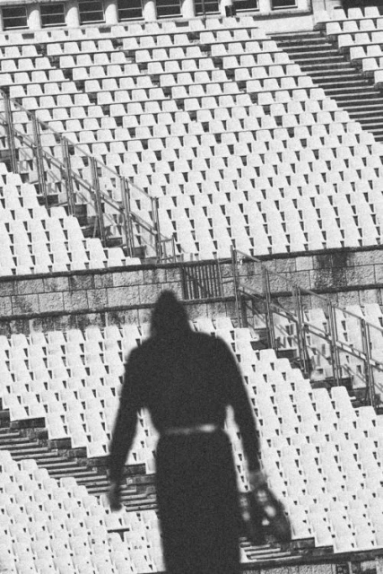 Monochromatic photograph featuring a silhouetted figure holding a glove in front of empty stadium seating. The image emphasizes stark contrast.