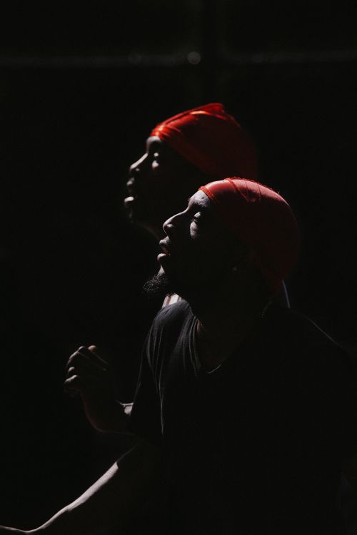 Profile view of two black adults wearing red headscarves against a dark background, illuminated by focused lighting.