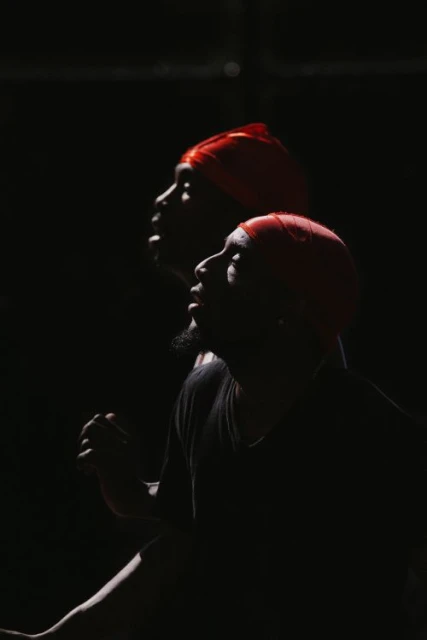 Profile view of two black adults wearing red headscarves against a dark background, illuminated by focused lighting.