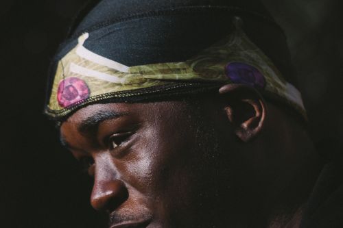 Close-up of a dark-skinned adult male wearing a black and patterned head covering, set against a dark, shadowed background.