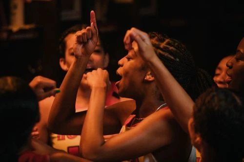 Excited group of young women with braids cheering together indoors, raising arms, and expressing joy, dark lighting, candid setting.