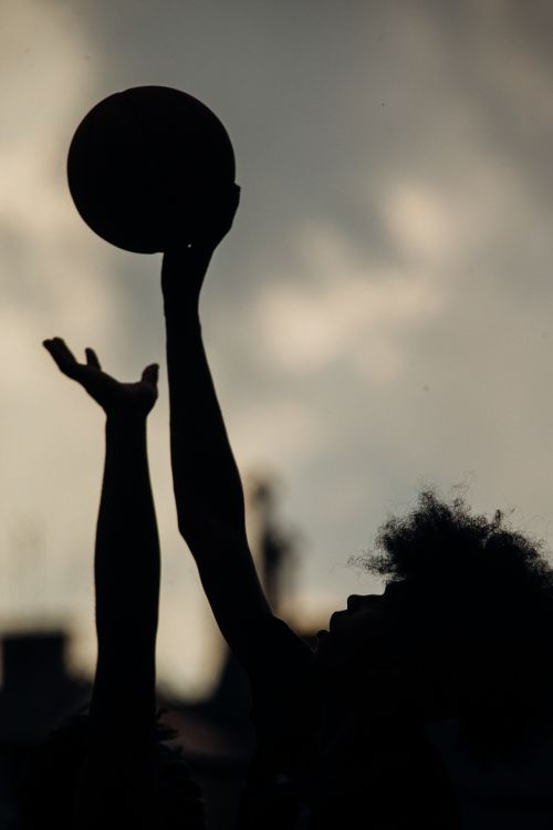 Silhouette of a man with afro hair holding a basketball with his arms up against an overcast sky. Focus on athlete and ball.
