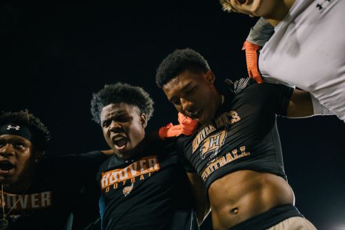 A close-up shot of four African-American men in football jerseys shouting and embracing, set against a dark background with stadium lighting.