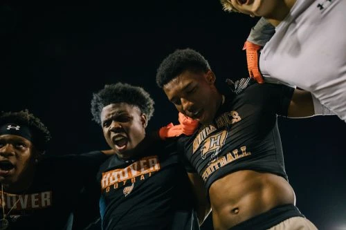 A close-up shot of four African-American men in football jerseys shouting and embracing, set against a dark background with stadium lighting.
