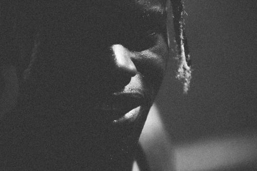 Monochrome close-up of a Black man's face in shadow, with a wet sheen on his skin and indistinct dark background, conveying a serious mood.