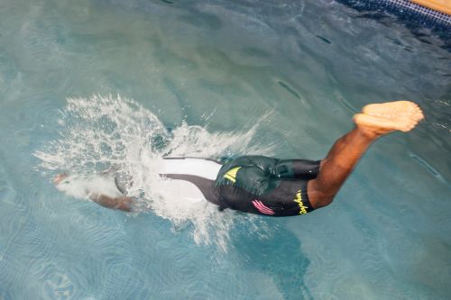 An adult dives into a blue-tiled swimming pool, kicking up a splash. The swimmer wears black and white swimwear. Clear water reflects the surrounding light.