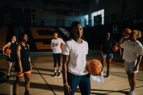 A group of young women athletes, in sports clothing, are indoors on a basketball court holding basketballs during a practice session.