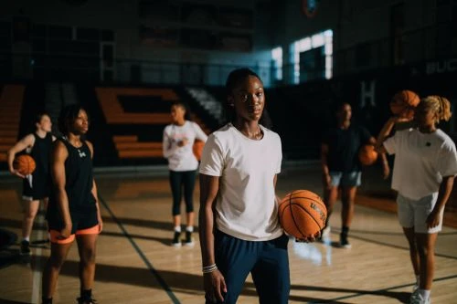 A group of young women athletes, in sports clothing, are indoors on a basketball court holding basketballs during a practice session.