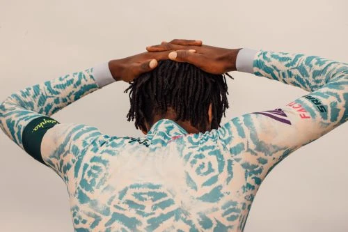 Back view of an adult male with dreadlocks wearing a patterned cycling jersey and holding his hands behind his head.