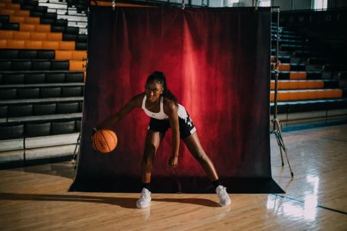 A female basketball player dribbling a basketball in front of a red studio backdrop inside a gymnasium, photographed by Alessandro Simonetti.