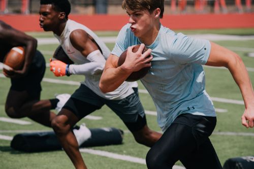 Three male football players in uniform run drills on a field on a sunny day with green grass and white lines, focus on the player in front.