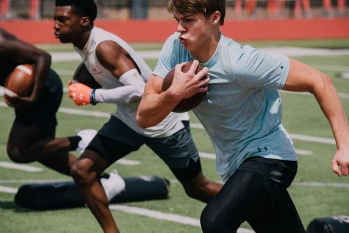 Three male football players in uniform run drills on a field on a sunny day with green grass and white lines, focus on the player in front.