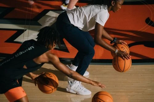 Two young Black women practice basketball indoors, dribbling orange basketballs on a wooden court with an athletic logo.
