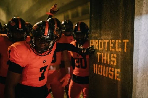 A group of football players in orange uniforms huddle near a concrete wall with the words 'Protect This House' spray-painted on it.