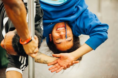Two men perform acrobatic moves on a pole indoors; one hangs upside down with a happy facial expression and the other holds the pole.