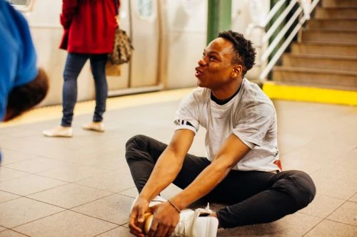A young adult male with short curly hair, wearing a gray tee shirt and dark pants, sits cross-legged on a subway platform.