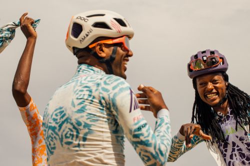 Three cyclists in patterned jerseys and helmets celebrate with wide smiles and raised arms against a bright, neutral sky. Action shot, sports theme.