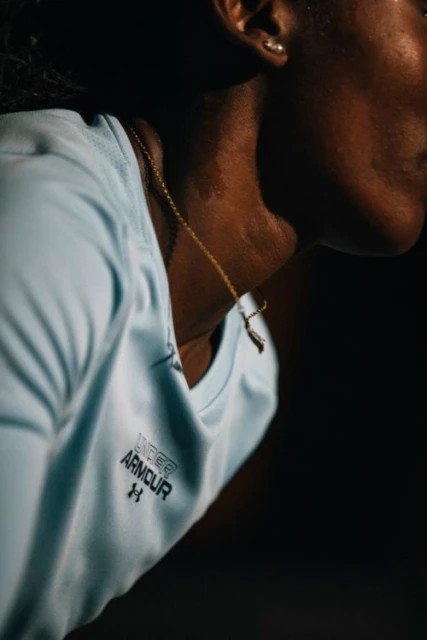 Profile of a woman wearing a light-blue Under Armour top and a gold chain, against a dark background, in an indoor environment.