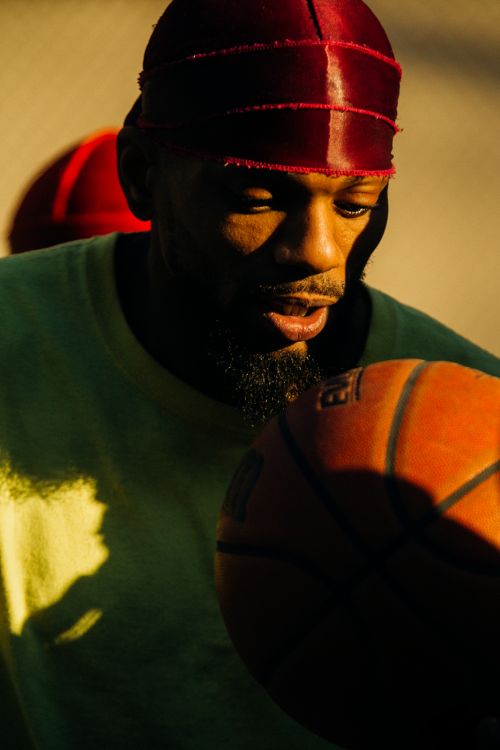 Man with beard and red head covering holds orange basketball in a close-up shot, bathed in soft, warm lighting.