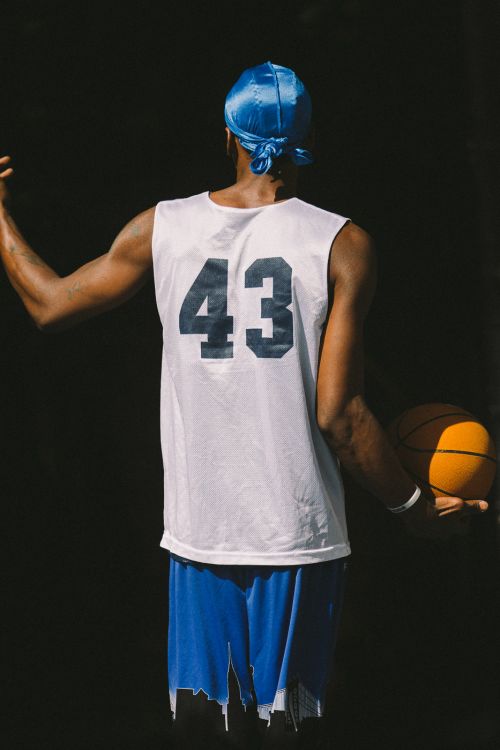 Back view of an African American male in a blue du-rag, white basketball jersey with '43', and blue shorts holding a basketball.