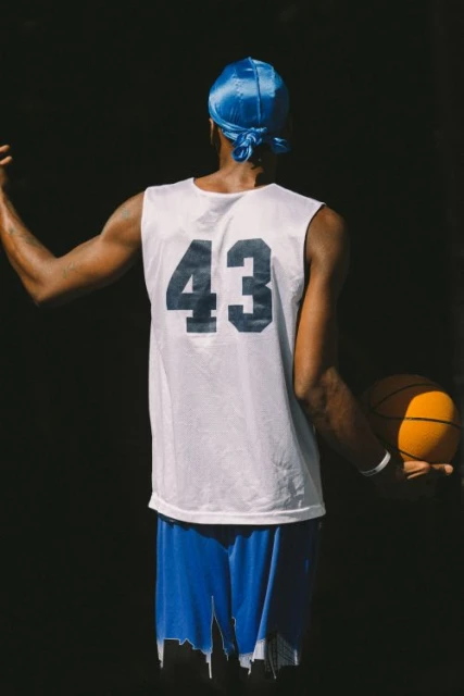 Back view of an African American male in a blue du-rag, white basketball jersey with '43', and blue shorts holding a basketball.