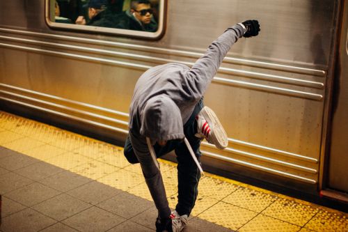 Adult bends over near subway train; wears hoodie, jeans, and sneakers. Subway platform with yellow tactile paving and gray tile visible.