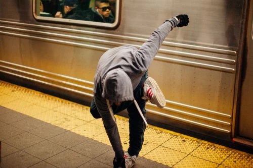 Adult bends over near subway train; wears hoodie, jeans, and sneakers. Subway platform with yellow tactile paving and gray tile visible.