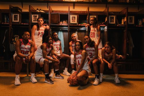 Group of diverse young women's basketball team in uniforms sitting together in a locker room with warm lighting and serious expressions.