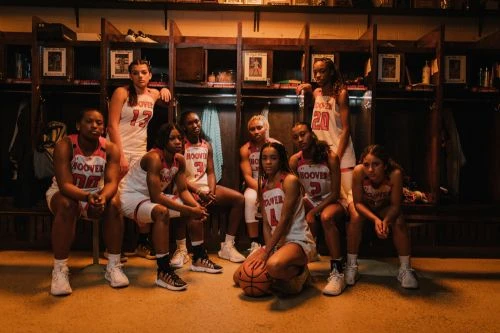 Group of diverse young women's basketball team in uniforms sitting together in a locker room with warm lighting and serious expressions.