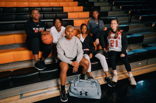 A group of six young adult women in sports apparel are sitting in a row on an indoor sports stadium bleacher, with a basketball.