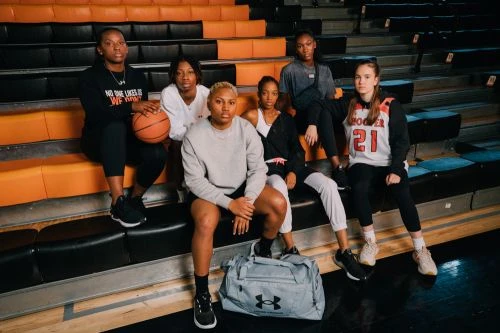 A group of six young adult women in sports apparel are sitting in a row on an indoor sports stadium bleacher, with a basketball.
