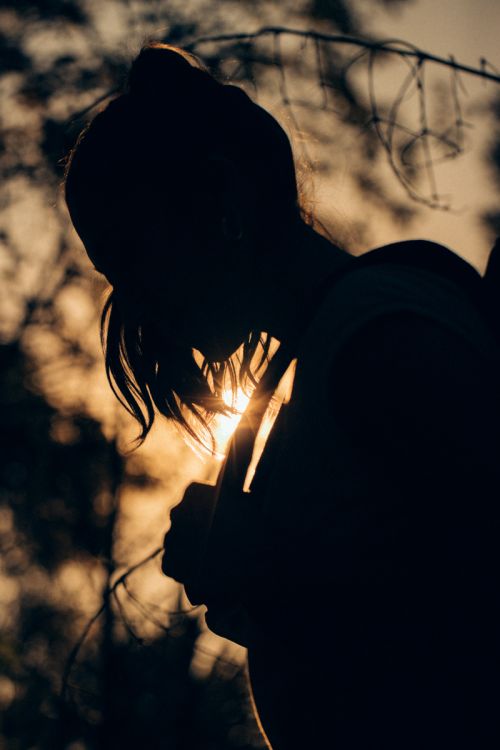 Silhouette of an adult female with dark hair against a bright sunlit sky with out-of-focus trees. Dark tonal mood.