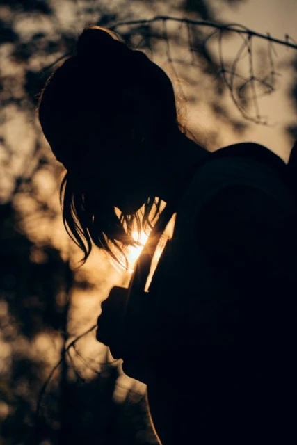 Silhouette of an adult female with dark hair against a bright sunlit sky with out-of-focus trees. Dark tonal mood.