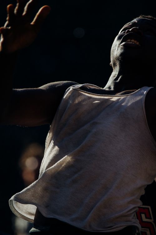 An African-American man with arms raised and a joyful expression, wearing a white tank top, against a dark, diffused background.