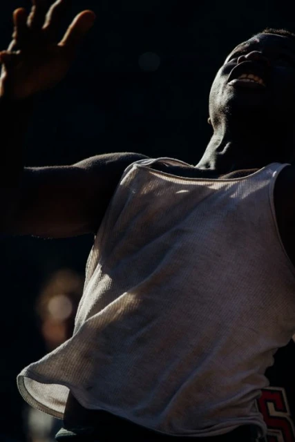 An African-American man with arms raised and a joyful expression, wearing a white tank top, against a dark, diffused background.