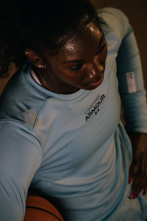 High-angle shot of an African-American woman in a light blue Under Armour shirt, dribbling a basketball in a dimly lit indoor setting.
