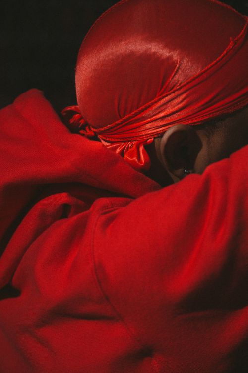 Close-up of an African American male wearing a red satin durag and a red hoodie in a dark, intimate setting with shallow depth of field.