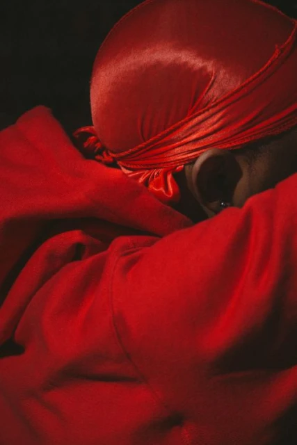 Close-up of an African American male wearing a red satin durag and a red hoodie in a dark, intimate setting with shallow depth of field.