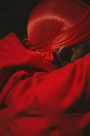 Close-up of an African American male wearing a red satin durag and a red hoodie in a dark, intimate setting with shallow depth of field.