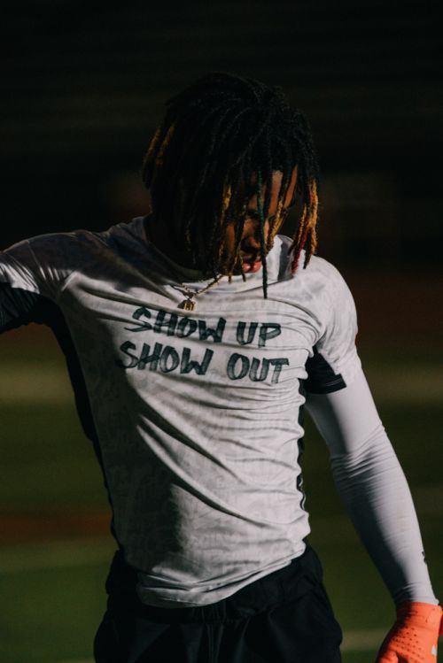 An athletic person in a long-sleeve shirt with the phrase 'Show Up Show Out' poses on a field at night under dim lighting.