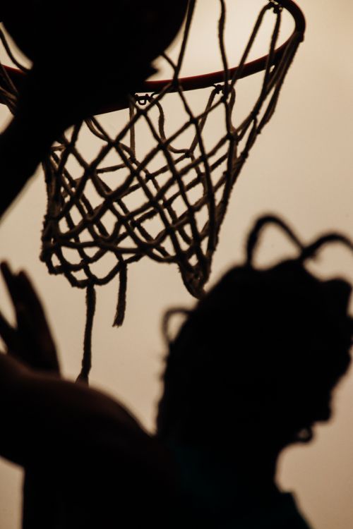 Silhouette of a person playing basketball with a basketball net and ball against a light background in an indoor setting.