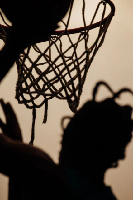 Silhouette of a person playing basketball with a basketball net and ball against a light background in an indoor setting.